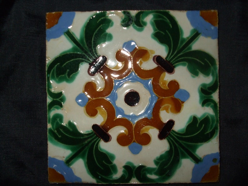 May include: A square ceramic tile with a white background and a colorful floral design. The design features green leaves, blue and brown swirls, and a black and white center.