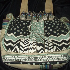 May include: A beige canvas tote bag with a design of three black, white, and green cats with blue eyes. The cats are sitting on a patterned background with a chevron design. The bag has a brown leather handle.