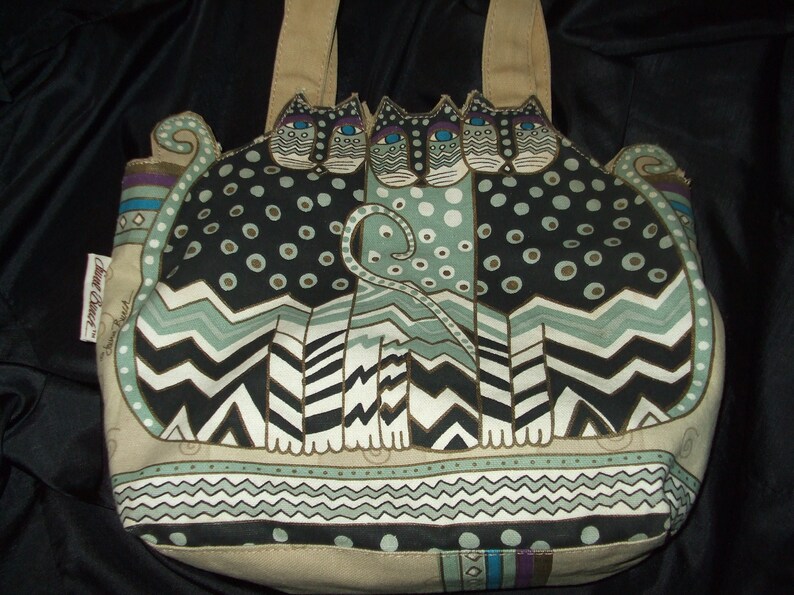 May include: A beige canvas tote bag with a design of three black, white, and green cats with blue eyes. The cats are sitting on a patterned background with a chevron design. The bag has a brown leather handle.