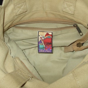 May include: A beige canvas tote bag with a zippered pocket and a small, colorful patch that reads "Anne Cole" inside.