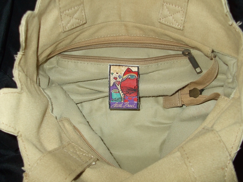 May include: A beige canvas tote bag with a zippered pocket and a small, colorful patch that reads "Anne Cole" inside.