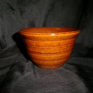 May include: A brown ceramic bowl with a glossy finish. The bowl has a rounded shape with horizontal ridges around the exterior. The interior is smooth and the bowl sits on a dark background.