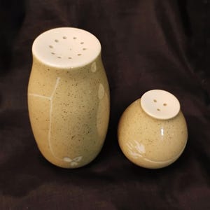 May include: A pair of ceramic salt and pepper shakers. The taller shaker is egg-shaped with a white top and a speckled tan body. The shorter shaker is round with a white top. Both have small holes for dispensing.