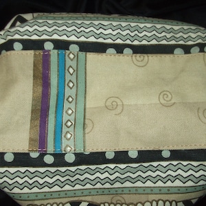 May include: A beige fabric tote bag with a black, white, and green geometric pattern. The bag has a large pocket on the front with a colorful striped design.