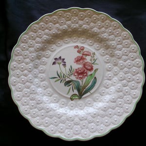 May include: White ceramic plate with a raised, embossed pattern of small flowers. The center of the plate features a floral design with pink, green, and purple flowers.