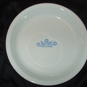May include: White pie plate with a blue floral design. The design features three blue flowers with green leaves.