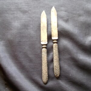 May include: Two antique silver butter knives with ornate floral patterned handles. The blades are slightly curved and show signs of age. The knives are displayed on a dark gray fabric.