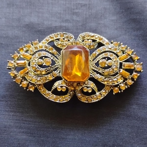 May include: An oval-shaped brooch featuring a large, central rectangular amber-colored stone surrounded by intricate gold-toned metal filigree work. The frame is embellished with numerous small, round, and baguette-cut amber crystals.