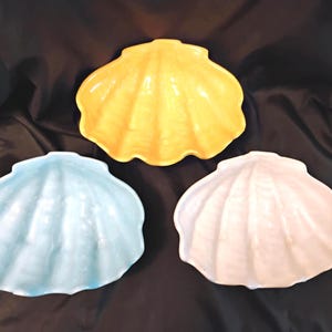 May include: Three ceramic seashell-shaped dishes in a variety of colors. The dishes are a light blue, pale yellow, and off-white. The dishes have a glossy finish and are arranged on a dark background.