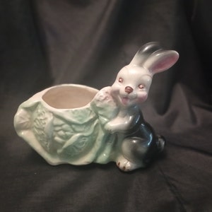 May include: A white ceramic planter shaped like a rabbit holding a green cabbage. The rabbit has black ears and a black tail.