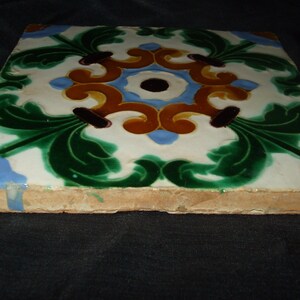 May include: A square ceramic tile with a decorative design featuring green, blue, yellow, and brown floral patterns. The tile has a white background and a slightly rough texture.