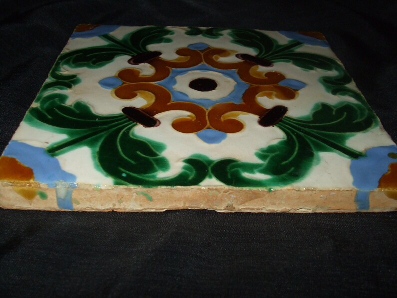 May include: A square ceramic tile with a decorative design featuring green, blue, yellow, and brown floral patterns. The tile has a white background and a slightly rough texture.