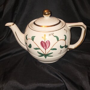 May include: A white teapot with a gold trim and floral design. The teapot has a round body and a curved spout. The flowers are pink, green, and yellow.
