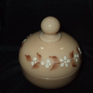 May include: A vintage glass jar with a lid, featuring a floral design in brown and white. The jar is a pale peach color.