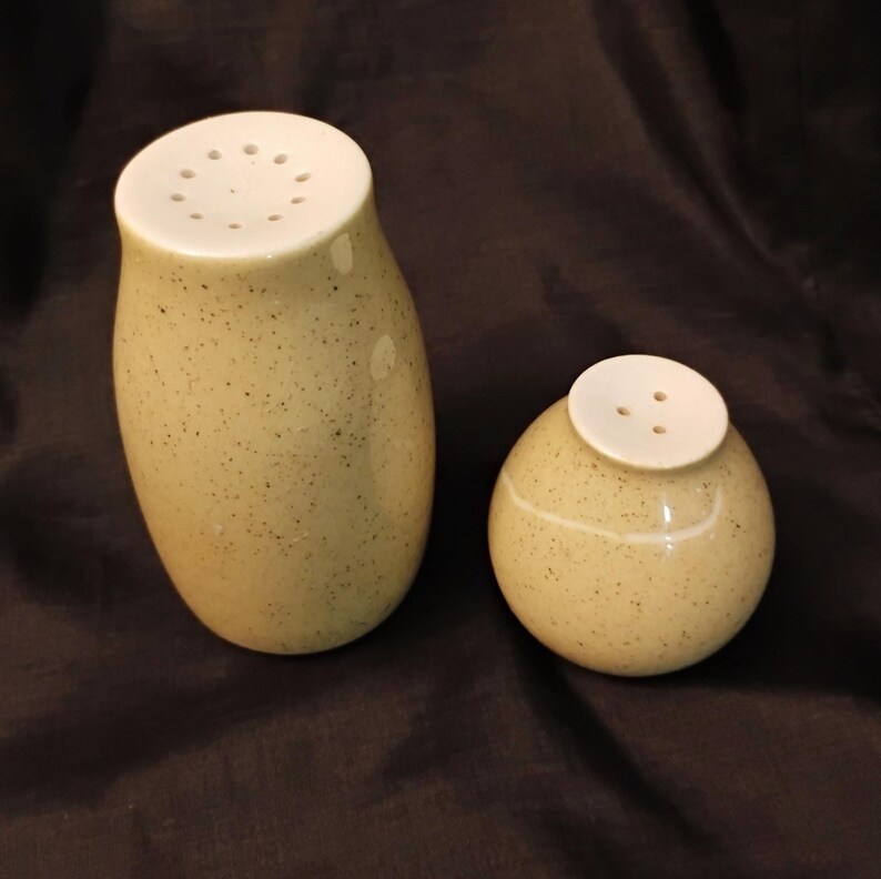 May include: A pair of speckled ceramic salt and pepper shakers. The taller shaker is oval-shaped, and the smaller one is round. Both have white tops with holes for dispensing. The shakers are a light beige color with dark speckles.