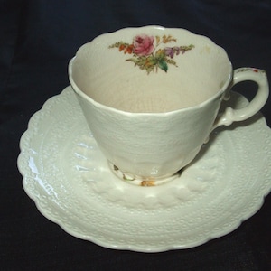May include: A white teacup and saucer set with a floral design. The cup has a pink rose and green leaves on the side. The saucer has a delicate lace pattern.