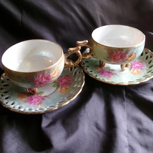 May include: Two vintage teacups and saucers with a floral design. The cups have gold trim and are decorated with pink and yellow roses on a green background. The saucers have a delicate lace pattern.