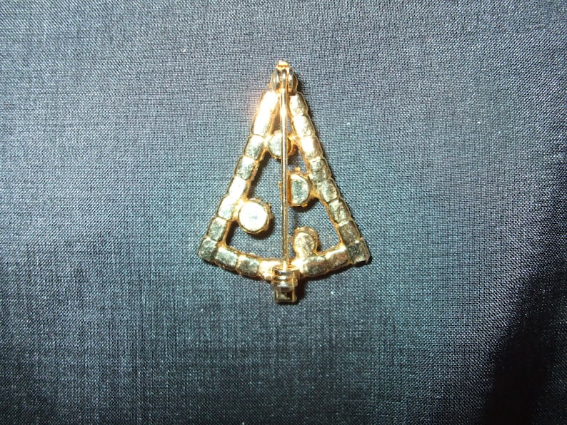 May include: Gold-tone Christmas tree brooch with clear rhinestones. The brooch is in the shape of a triangle and has a pin back.