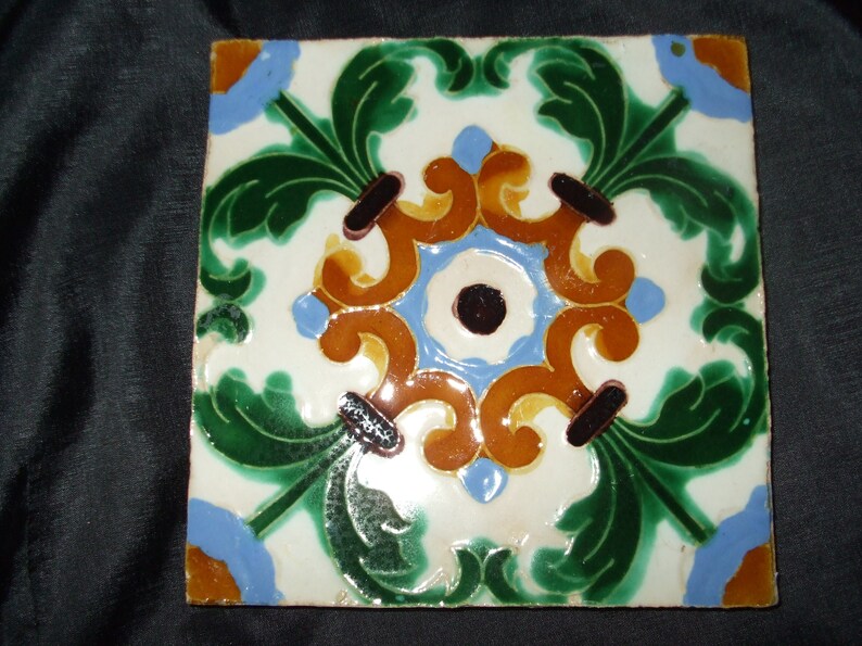 May include: A square ceramic tile with a decorative design featuring green, blue, brown, and yellow floral motifs. The tile has a white background and a glossy finish.
