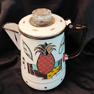 May include: White enamel coffee pot with a pineapple design. The pot features a clear glass lid, a black handle with colorful accents, and gold trim. The pineapple is red and brown, with a yellow and blue background.