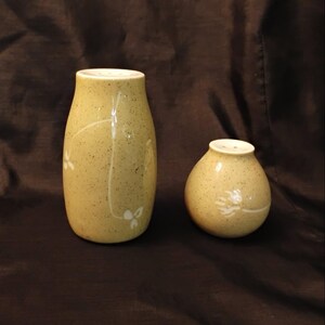 May include: A pair of ceramic salt and pepper shakers. The shakers are a speckled, light yellow-green color with white floral designs. The taller shaker is approximately 4 inches tall, and the shorter shaker is about 3 inches tall.