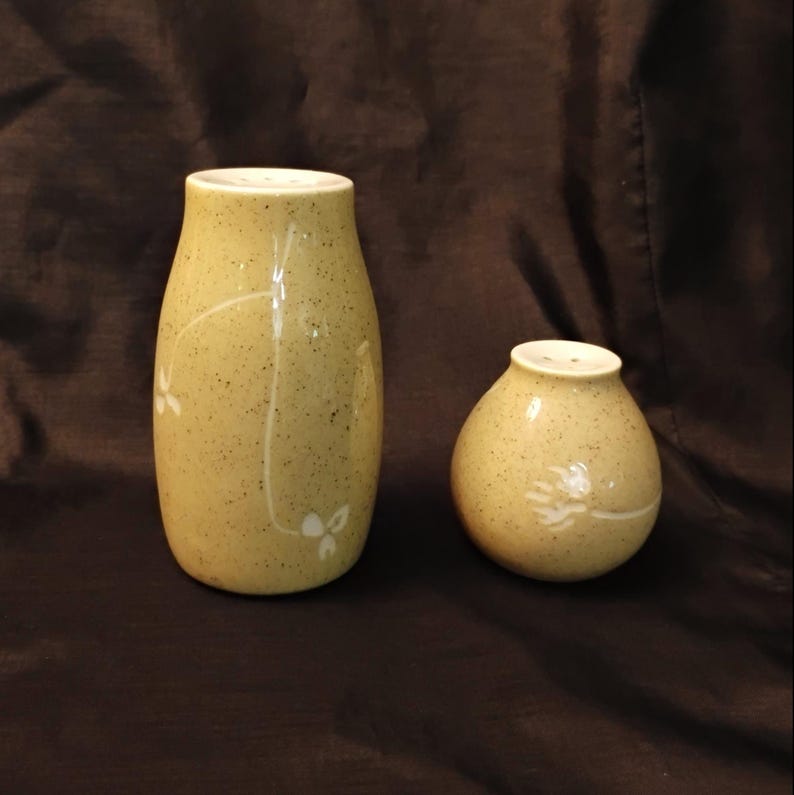 May include: A pair of ceramic salt and pepper shakers. The shakers are a speckled, light yellow-green color with white floral designs. The taller shaker is approximately 4 inches tall, and the shorter shaker is about 3 inches tall.