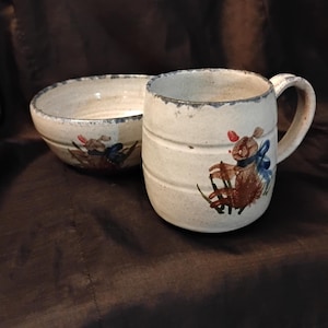 May include: A ceramic mug and a matching bowl with a speckled off-white glaze. Both pieces feature a hand-painted brown rabbit illustration with a blue bow and red accents. The mug has a cylindrical shape with horizontal ridges and a side handle.