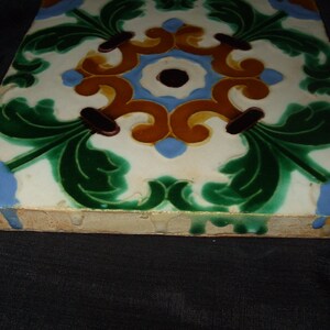 May include: A square ceramic tile with a white background and a colorful floral design. The design features green leaves, brown stems, and yellow and blue swirls.
