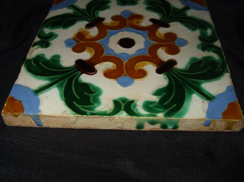 May include: A square ceramic tile with a white background and a colorful floral design. The design features green leaves, brown stems, and yellow and blue swirls.