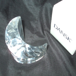 May include: A clear glass crescent moon shaped candle holder with the word "Dansk" printed on the white box it is sitting on.