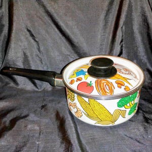 May include: A white saucepan with a black handle and lid. The lid and the body of the pan are decorated with colorful illustrations of vegetables, including corn, tomatoes, and mushrooms. The pan is designed for cooking.