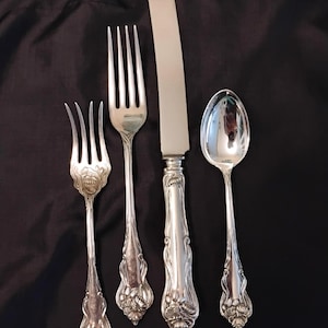 May include: Set of four antique silver-colored flatware pieces: a knife, a spoon, and two forks. The handles feature an ornate floral design. The flatware is arranged on a black background, showcasing the reflective quality of the metal.