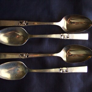 May include: Four silver spoons with ornate handles. The spoons are arranged in a row, with the handles facing the viewer. The spoons are all the same size and shape.