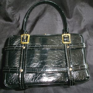 May include: A black patent leather handbag with gold-toned hardware and a crocodile-embossed design. The bag has a top handle and a structured shape.