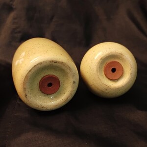May include: Two ceramic salt and pepper shakers with a speckled, pale yellow glaze. Each shaker has a round, dark brown stopper. The shakers are egg-shaped and have a small white floral design on the side.