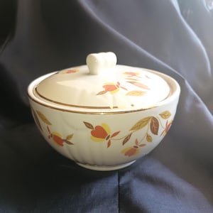 May include: A white ceramic bowl with a lid, featuring a floral pattern with orange, yellow, and brown leaves. The bowl has a gold trim around the rim.