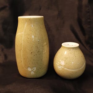 May include: A pair of speckled ceramic vases in a muted yellow-green hue. The taller vase has a slender, elongated shape with a floral design. The smaller, round vase has a similar floral design and a series of holes at the top. Both vases have a glossy finish.