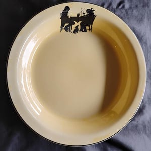 May include: A round, cream-colored enamel bowl with a dark rim. The interior features a black silhouette graphic near the top edge depicting two figures seated at a table with a small dog standing between them, rendered in a simple, flat style.
