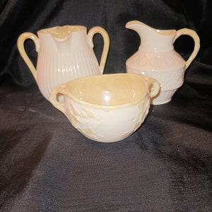 May include: A set of three cream-colored ceramic pieces with yellow accents. The pieces include a creamer, a sugar bowl, and a pitcher. All three pieces have a floral design.