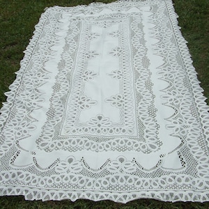 May include: White lace tablecloth with a delicate floral pattern and a scalloped edge. The tablecloth is laid out on a green grassy surface.