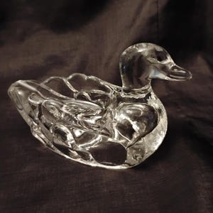 May include: A clear glass figurine shaped like a duck, featuring a smooth, reflective surface and detailed molded wings. The duck is depicted in a resting position with its head turned slightly to the side, showcasing a solid, transparent body.