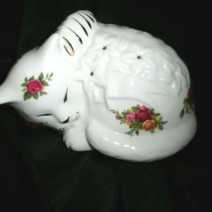 May include: A white porcelain figurine of a sleeping cat with a floral design. The cat is curled up with its head resting on its paws. The floral design features red, yellow, and green roses.