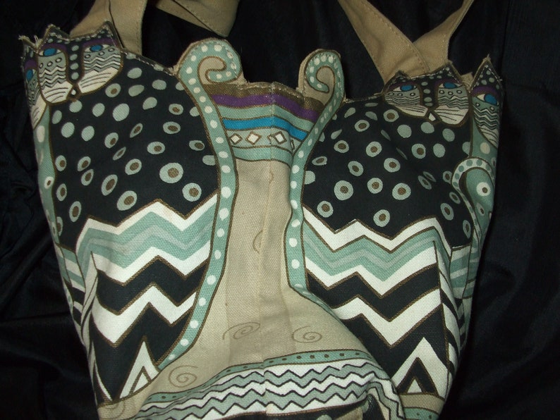 May include: A beige tote bag with a black, white, and green cat design. The cats have a geometric pattern with circles, zigzags, and swirls.