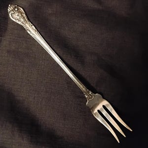 May include: A silver-toned metal cocktail fork featuring an elongated handle with intricate embossed floral and scrollwork detailing at the top and a three-pronged head. The utensil has a polished finish and a slender, tapered design.