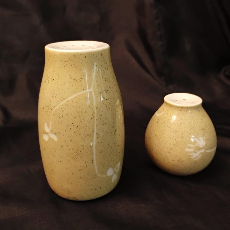 May include: A pair of ceramic salt and pepper shakers. The shakers are a speckled, light yellow-green color with white floral designs. The taller shaker is cylindrical, and the shorter one is round.