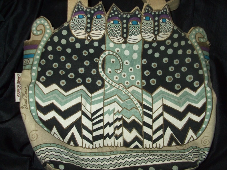 May include: A beige tote bag with a black, white, and green pattern of three stylized cats. The bag has a brown leather handle and a label that reads "Susan Branch".