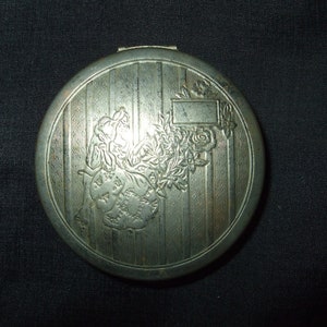 May include: Silver compact with an engraved design of a woman holding flowers. The compact has a striped pattern and a blank rectangular space for engraving.