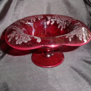 May include: A ruby red glass bowl with a footed base and a scalloped rim. The bowl is decorated with an etched floral design.