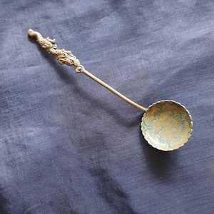 May include: A brass spoon with a long, slender handle featuring a sculpted dragon figure at the top. The bowl of the spoon is circular with a scalloped edge and is decorated with an intricate, engraved floral pattern across its surface.