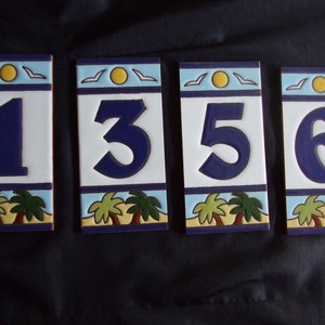 May include: Four ceramic house numbers with a beach theme. The numbers are 1, 3, 5, and 6. Each number is on a white background with a blue border and a yellow sun with white birds flying above. The bottom of each number has a brown sand beach with two green palm trees.